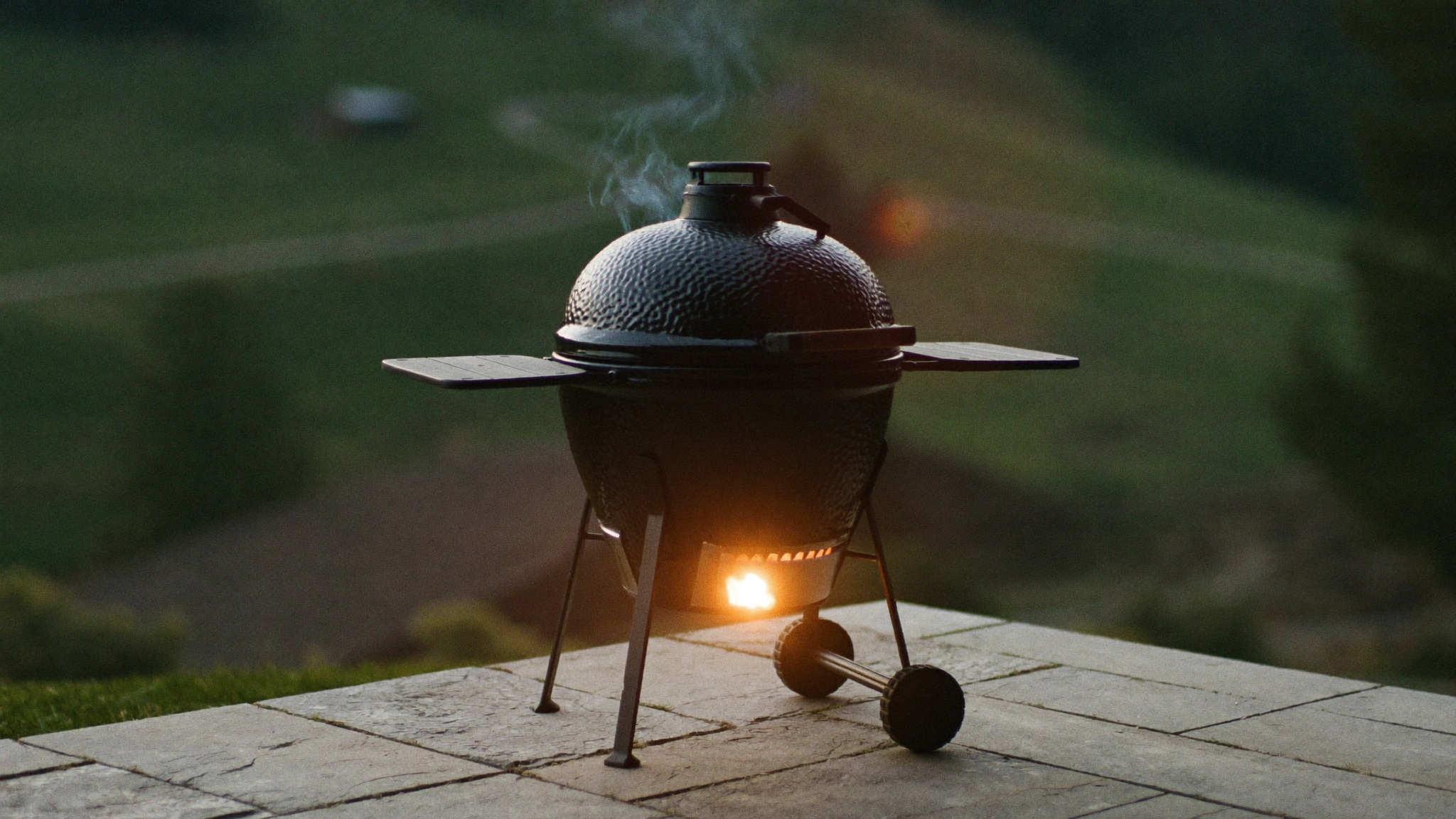 Geschmiedeter Kamado-Grill auf einer stillen Steinterrasse in der Abenddämmerung, Glut schimmert durch die Lüftungsschlitze, Rauch steigt auf